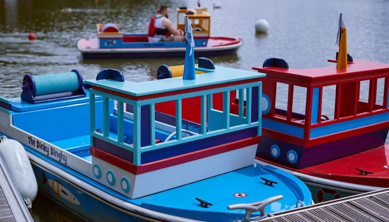 Colorful miniature boats float and dock on a calm lake; in the background, one boat is being ridden near buoys. Text: "The Dinky Dinghy".
