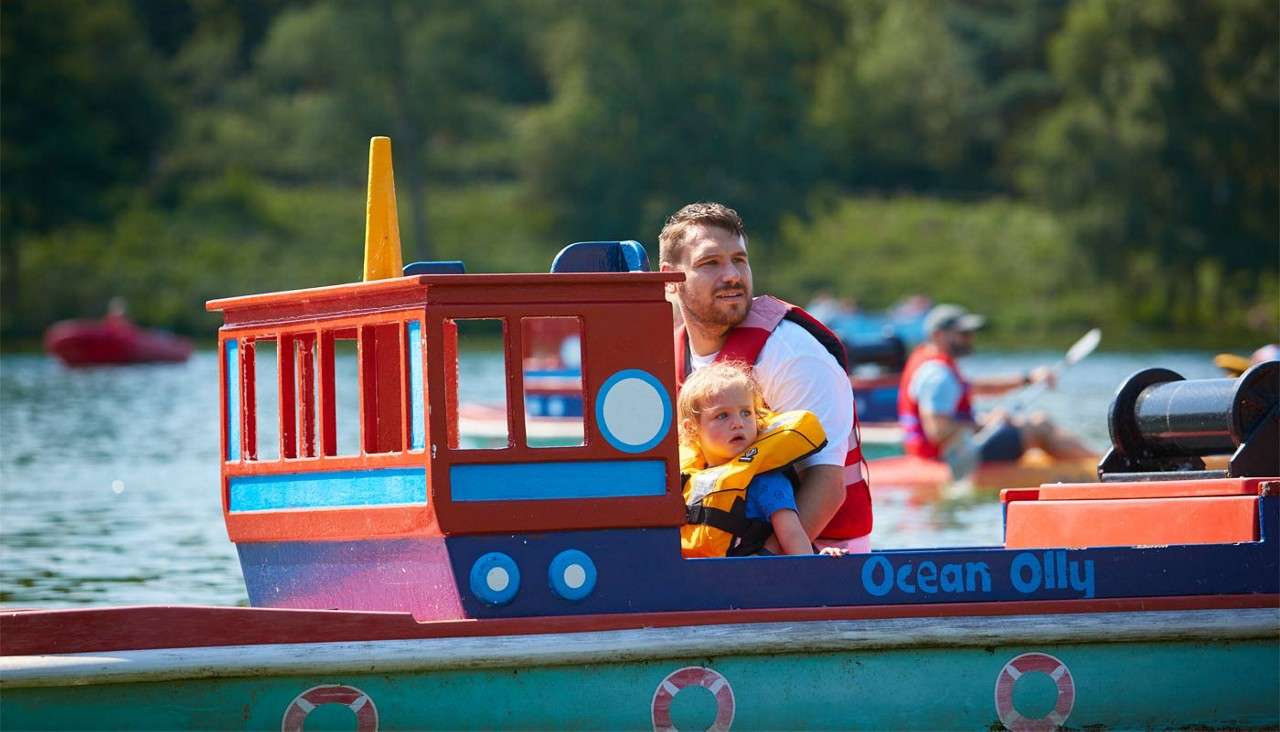 Adult and child ride a colorful toy boat, "Ocean Olly," wearing life jackets, navigating a calm lake, while other paddlers in red vests move in the background near forested shore.