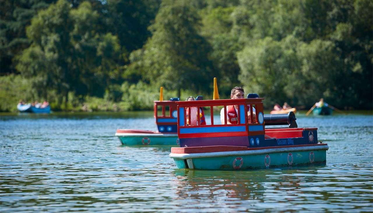 Two small red-and-blue toy boats carry riders steering slowly across a calm lake; wooded shoreline and other rowboats appear in the background. Text on hull: "Ocean ..." (remainder unreadable).