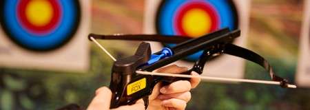 Pistol crossbow aims a blue bolt toward two concentric archery targets, string drawn taut, held by a hand in an indoor range with a blurred background.