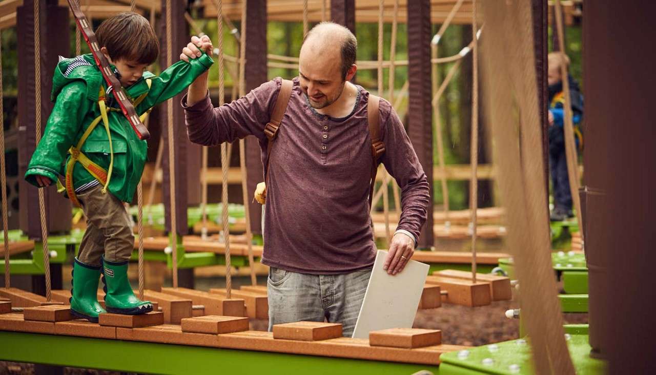 Child crosses a suspended wooden plank bridge, holding an adult’s hand, while wearing a harness and rain boots; adult steadies him in a forest ropes course with ropes and platforms.