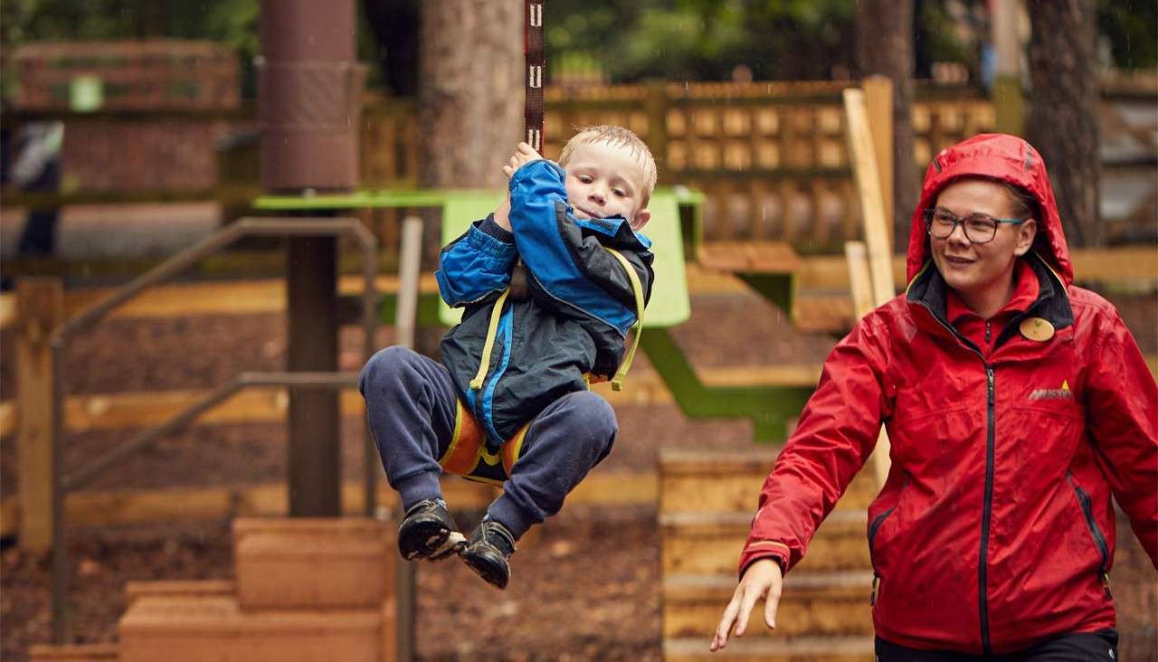 Child grips a zip-line handle, gliding forward; an adult in a red raincoat reaches out, supervising in a wooden outdoor playground surrounded by trees on a rainy day.