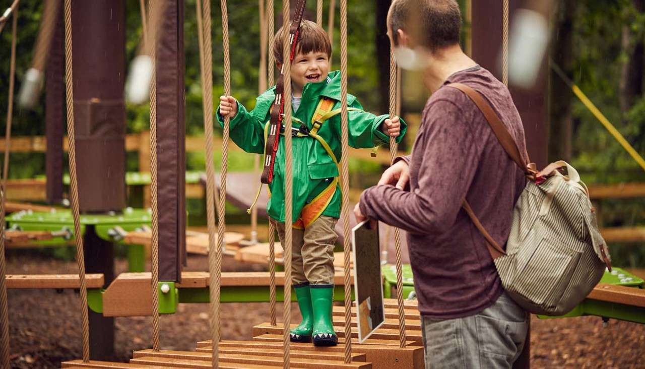 Child wearing harness crosses a suspended rope bridge, gripping ropes and smiling; adult supervisor stands nearby with backpack, guiding. Context: wooded outdoor adventure park with platforms and safety lines.