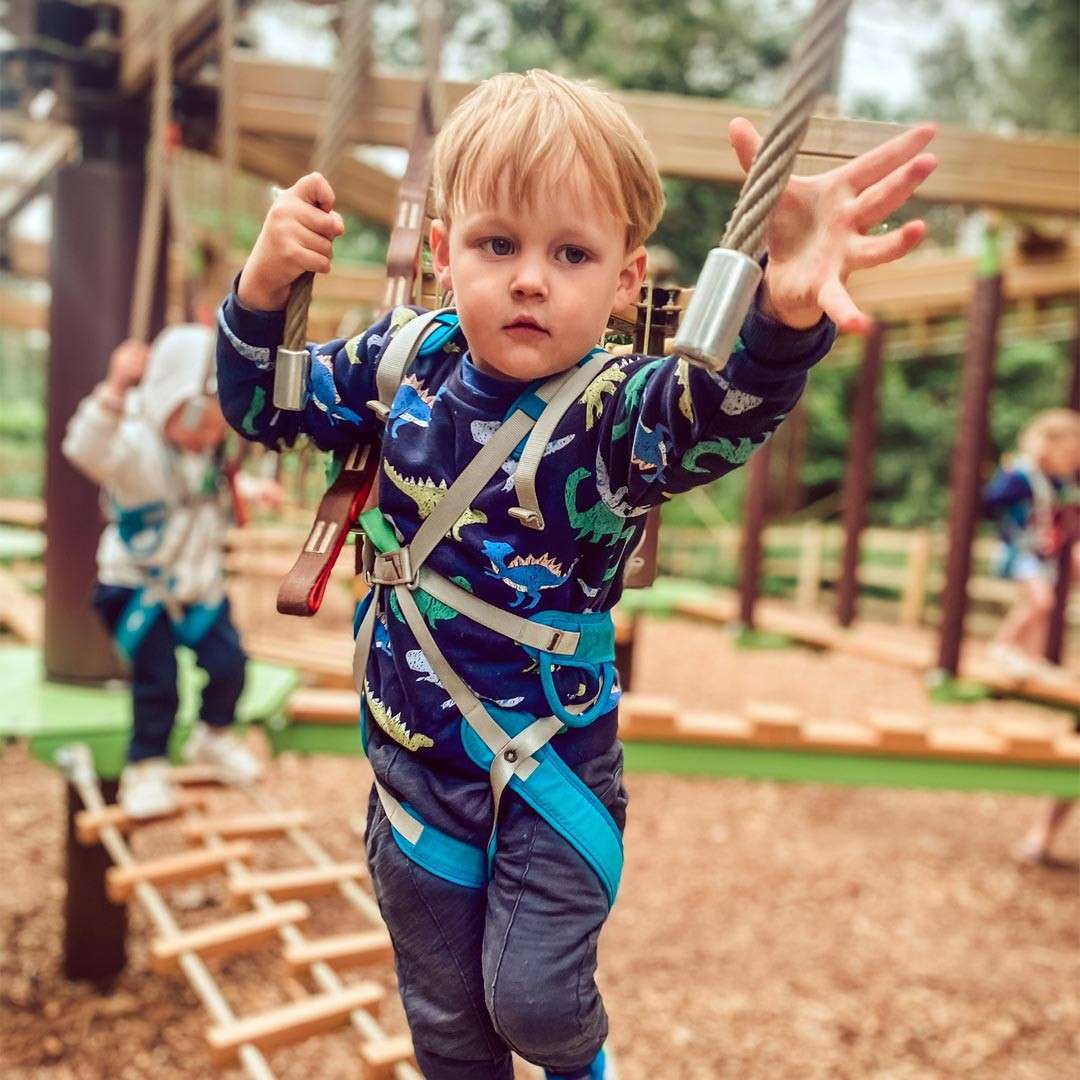 Child—secured in a harness—grips two ropes and reaches forward, balancing on a suspended obstacle. Background shows other children on a wooden ropes course in an outdoor play area.