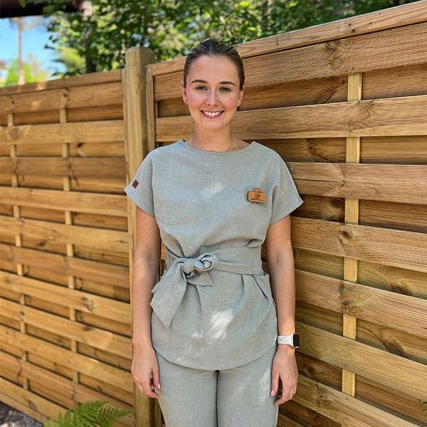 Person smiles while standing relaxed, hands by sides, wearing light-gray uniform with tied waist and watch. Context: sunny outdoor setting beside a wooden slat fence with green foliage.