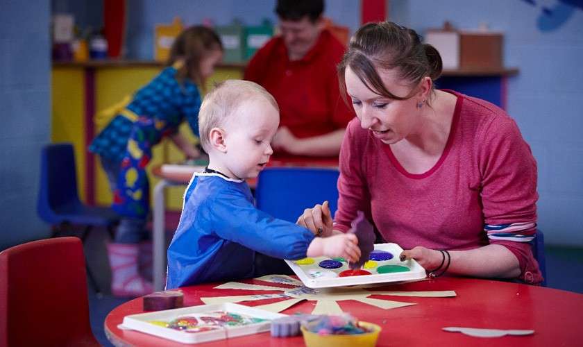 Child and caregiver finger-paint using a palette, dabbing colors onto paper. They sit at a red table in a colorful classroom, with other children and art supplies in the background.