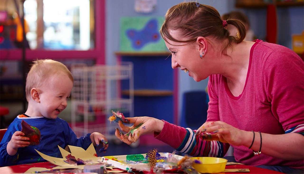 Child and adult finger-paint and laugh, smearing colorful paint on paper and hands, at a craft table in a classroom with supplies, shelves, and a butterfly picture in the background.