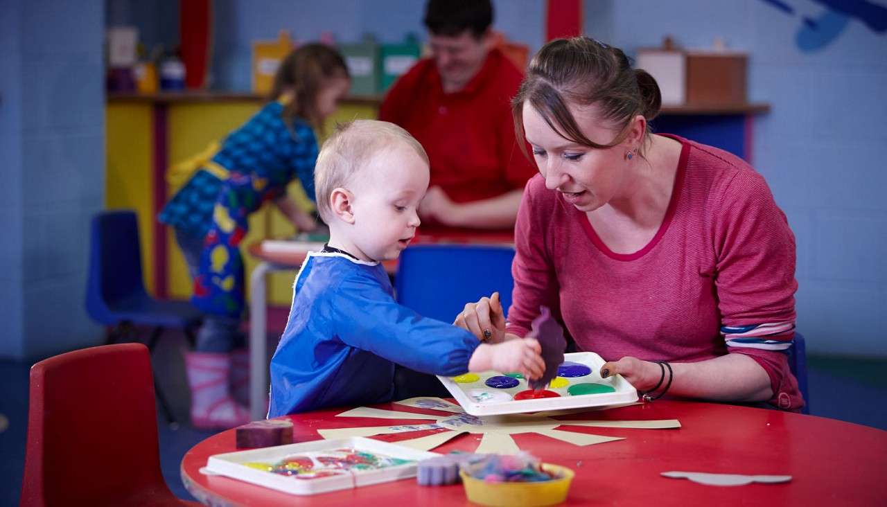 Child and caregiver mix bright paints on a palette, guiding a brush over paper in a colorful classroom with tables, craft supplies, and other children and adults working in background.