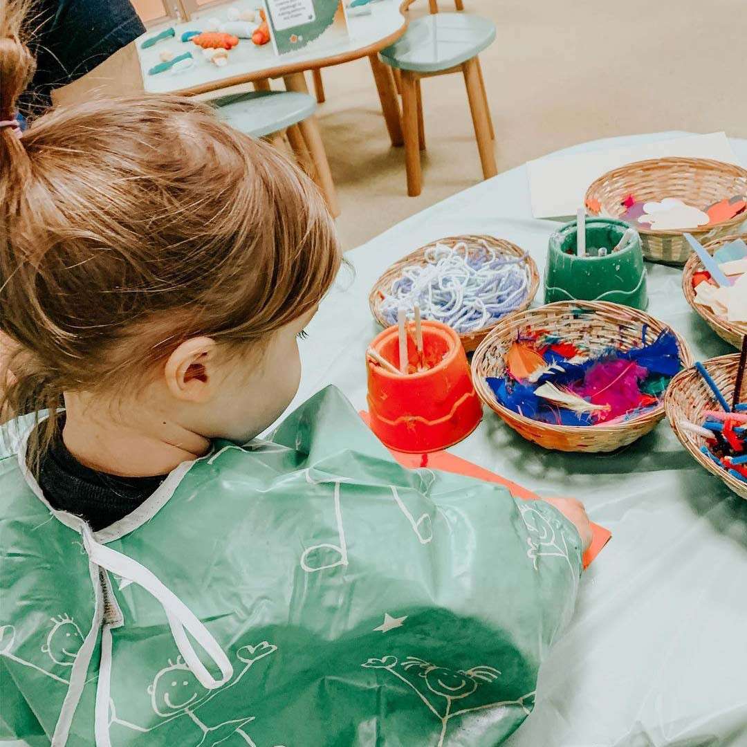 Child arranges craft materials, gluing colorful feathers and yarn on paper, while wearing a green smock; baskets of supplies and stools around suggest a children’s art station in a classroom.