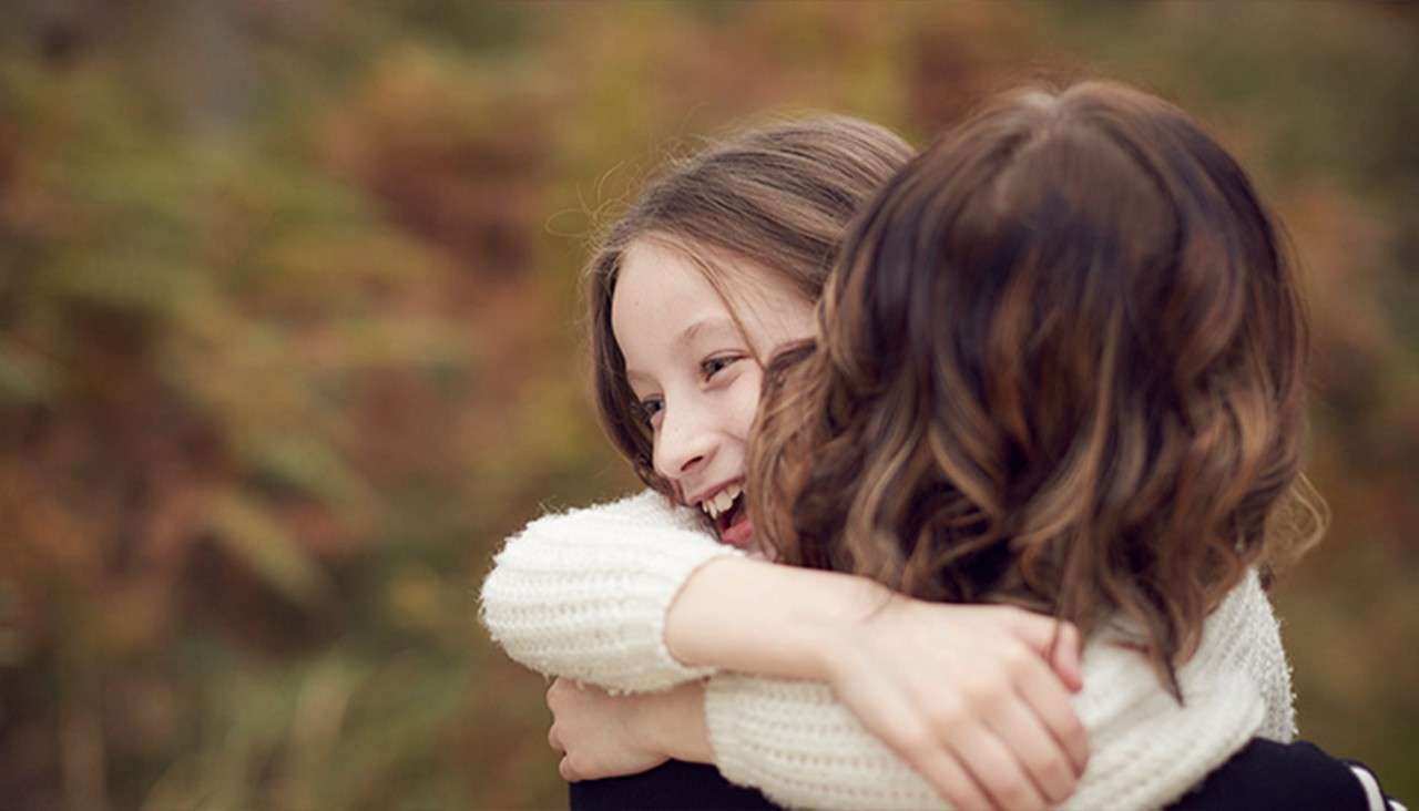 Smiling child hugs an adult tightly, arms wrapped around shoulders, face joyful. They stand outdoors amid soft-focus autumn foliage, both wearing cozy knit sweaters.