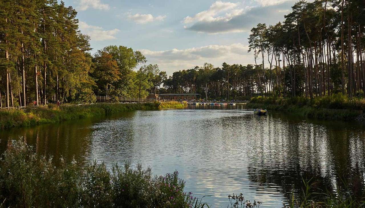 Calm river reflects tall pines; small paddle boats drift near a distant boathouse. Dense forest and grassy banks surround, while few people stroll along a path under cloudy sky.