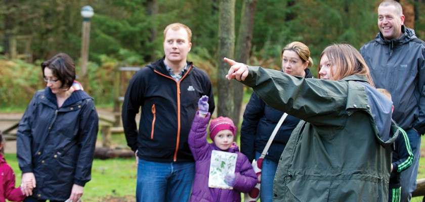 Guide points ahead, leading a small group; adults and children in jackets listen, one child raises a hand holding paper, in a forest clearing with trees and simple wooden play structures.