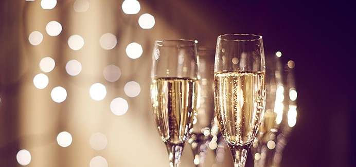 Champagne flutes stand filled with bubbly wine, bubbles rising. In the background, warm bokeh string lights glow softly, suggesting a festive, evening celebration.