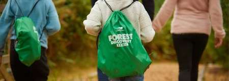 Child wearing a green drawstring backpack walks with companions along a forest path; backpack text reads: “FOREST DISCOVERY TRAIL.”