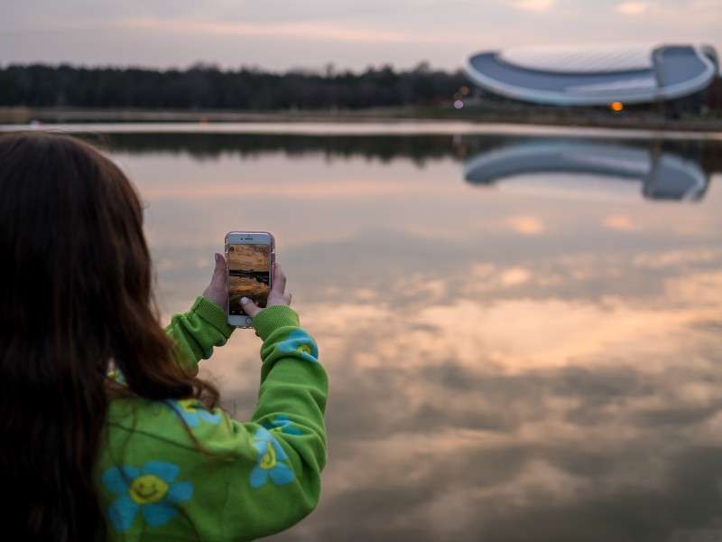 Person holding a smartphone photographs a calm lake at sunset, capturing reflections. Context: still water mirrors clouds, distant treeline, and a curved modern building across the shore.