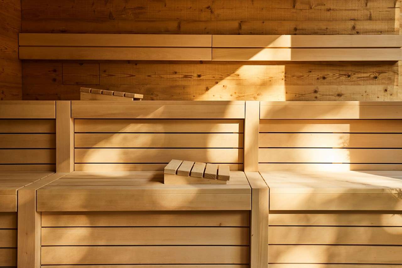 Wooden sauna benches sit in tiers, catching angled sunlight; two slatted headrests rest on the seats; warm paneled walls and shadows frame a quiet, empty sauna interior.