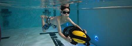 Child in swim goggles rides a yellow underwater scooter, gliding along the pool floor. Lane markings and blue lights appear nearby in a clear indoor swimming pool.