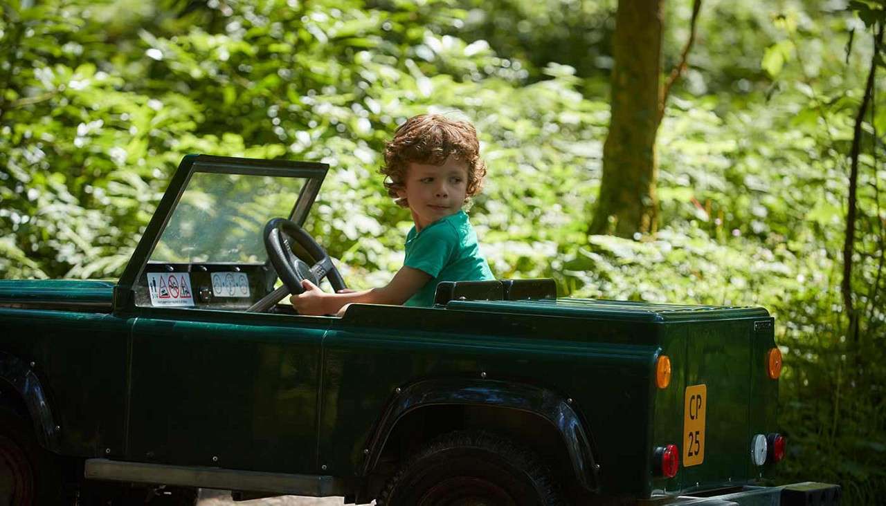 Child drives a small green toy jeep, turning the steering wheel and looking back; surrounding sunlit forest path with lush greenery. License plate reads CP 25.