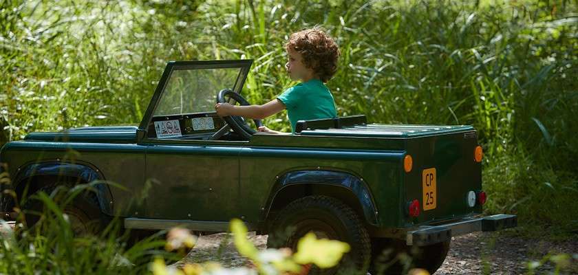 Child drives a small green toy off-road car on a gravel path, surrounded by tall grass and foliage; dashboard shows stickers; rear license-style sticker reads “CP 25.”