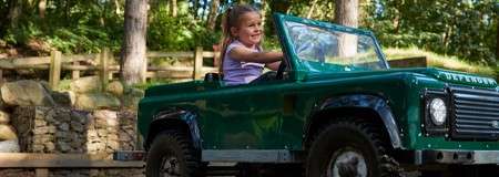 Child drives a green miniature Land Rover, smiling while steering. The car’s front reads “DEFENDER.” Surroundings: wooded park with sunlight, wooden fence, stone wall, and dirt path.