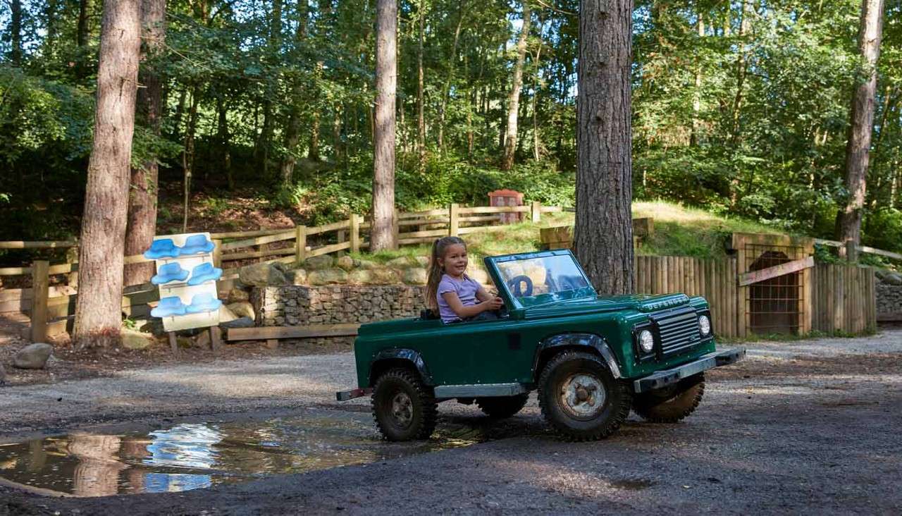 Child in a small green off-road toy car steers through a puddle; the car’s front reads DEFENDER. Surrounding trees, wooden fences, and gravel paths form a wooded park setting.
