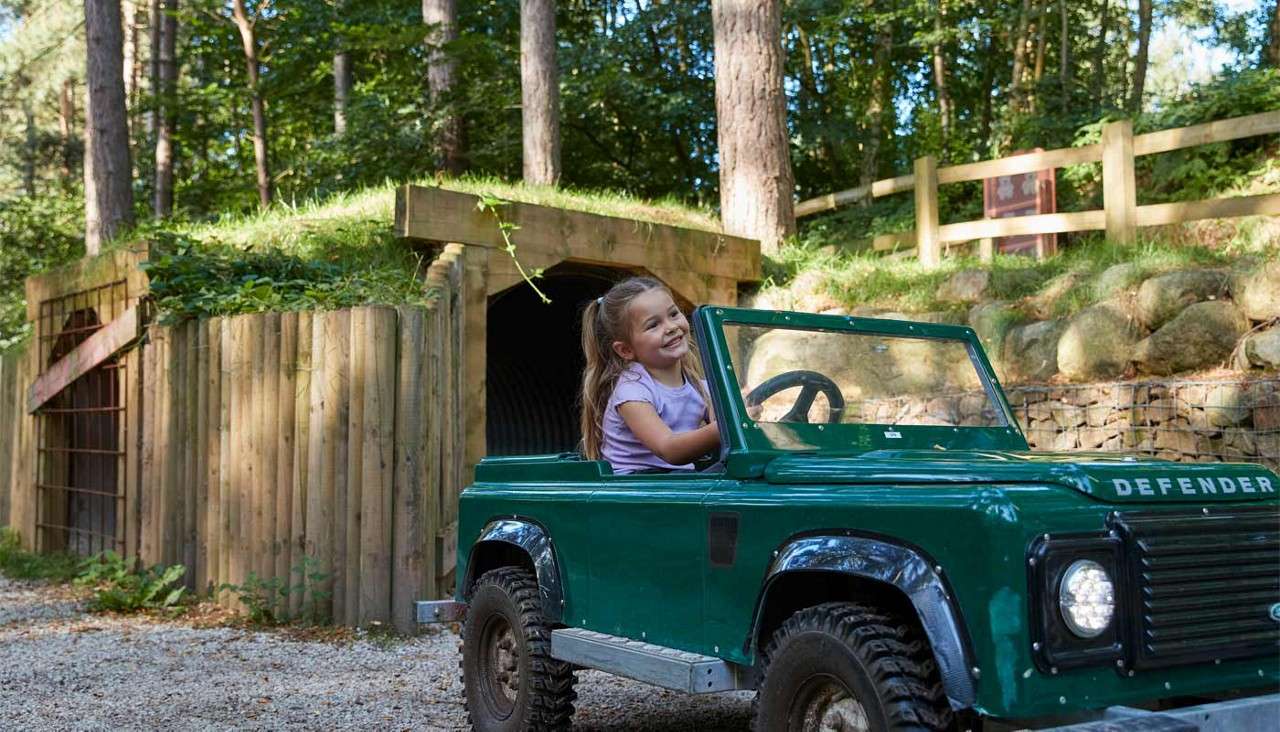 Child drives a small green open-top toy vehicle along a gravel path; wooded park setting with a timber tunnel entrance and fence behind. Visible text: DEFENDER.