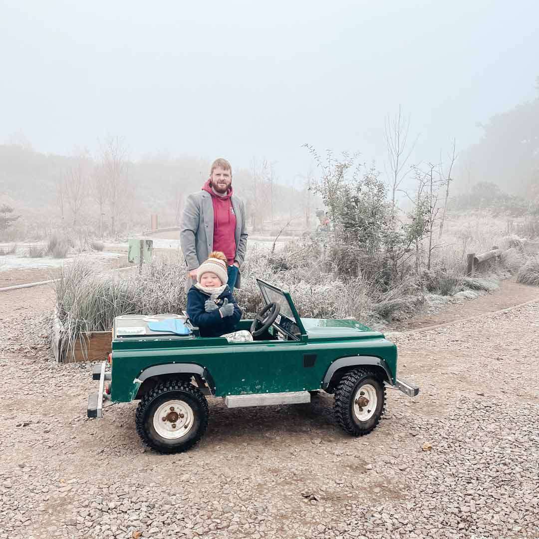 Child in a small green toy jeep sits at the wheel, smiling and giving a thumbs-up; adult stands behind, supervising on a frosty, foggy gravel path amid wintery shrubs.