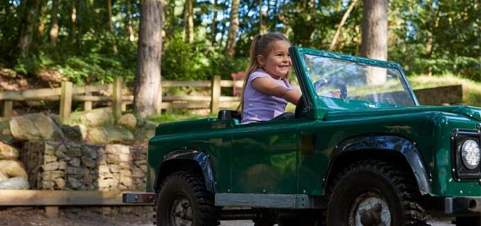 Child drives a small green toy jeep, smiling and steering. The vehicle moves along a dirt path in a wooded park, near a low stone wall and wooden fence.