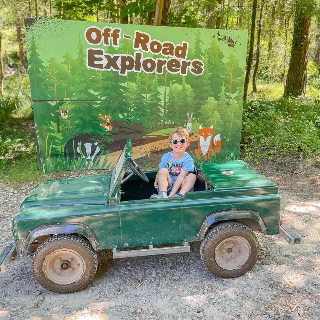Child sits in a small green toy jeep, smiling as if driving, in front of a forest-themed sign reading “Off-Road Explorers,” along a sunlit wooded trail.