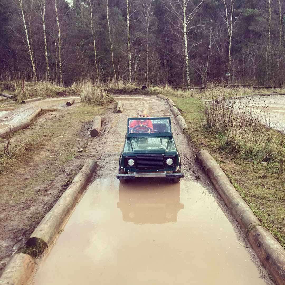 Small green toy jeep driven by a child splashes through a wide muddy puddle, following a log-lined dirt track in a sparsely grassy, wooded area under an overcast sky.