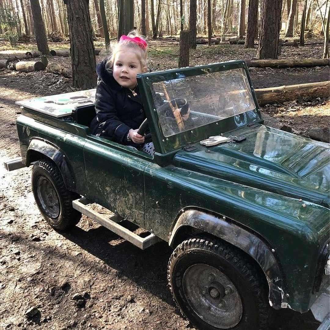 Child drives a small green toy jeep along a muddy forest path, seated behind a plastic windshield with hands on the steering wheel; tall trees and scattered logs surround.