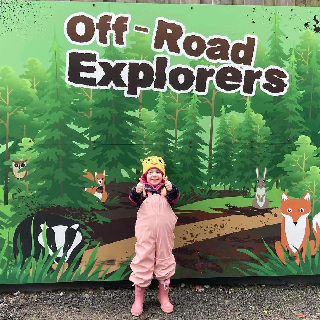 Child smiles and gives two thumbs-up, standing in pink rain gear and boots before a green forest-themed sign with cartoon animals. Text reads: Off-Road Explorers.