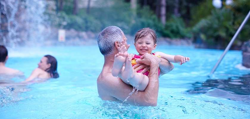 Adult man lifts a smiling toddler, splashing water, in a bright blue outdoor pool; waterfall and steps nearby; other swimmers and lush greenery in the background.
