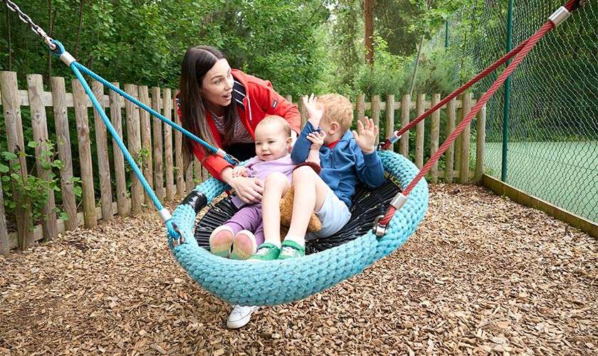 Two children sit in a large blue rope basket swing while an adult steadies them, outdoors on a playground with wood-chip ground, wooden fence, netting, and surrounding green trees.