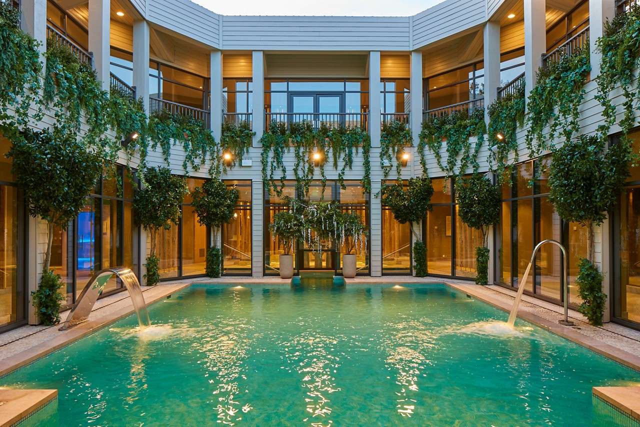 Pool sends arcing jets into turquoise water, set in a circular courtyard where glass walls, balconies, warm lights, hanging vines, and potted trees surround the tranquil, modern space.