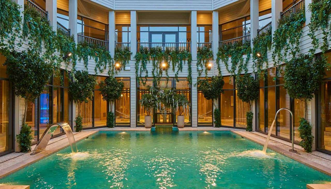 Swimming pool sends arcing water streams; turquoise surface ripples. Glass-walled courtyard surrounds two levels of balconies, warm lights, hanging vines, and potted trees, forming a serene indoor oasis.