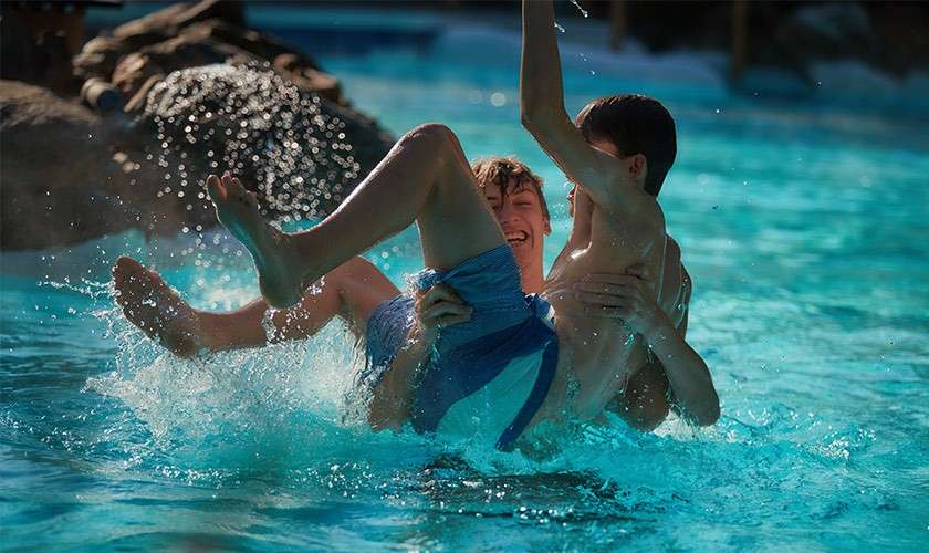 Two children play as one lifts the other, water splashing; turquoise swimming pool with sunlit ripples and a small cascading waterfall over rocks in the background.