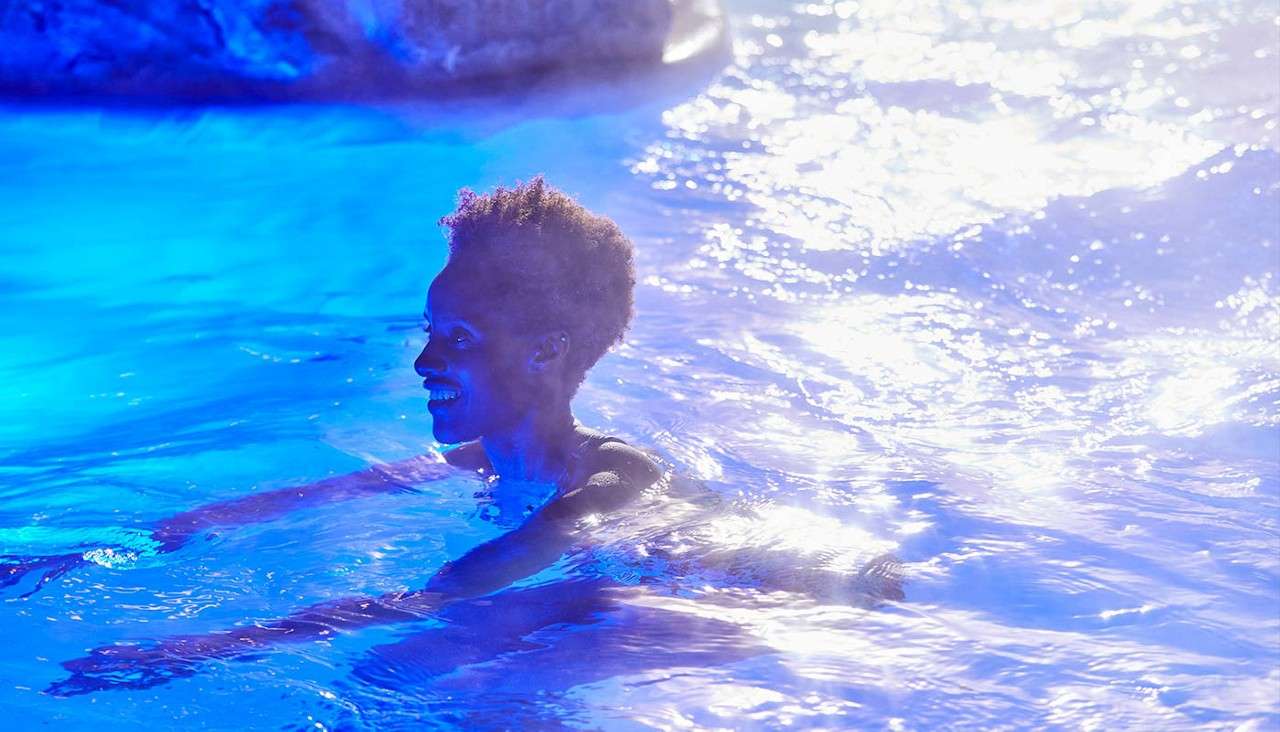 Swimmer smiles while gliding through illuminated blue water, set in a pool at night with rippling reflections and a rocky edge in the background.