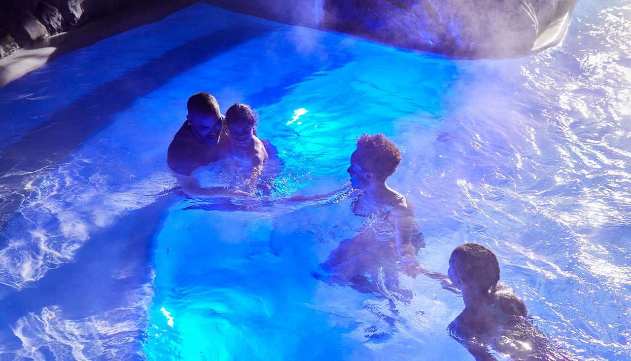 Four people wade and talk, holding hands in shallow water; blue pool lights glow beneath them as steam rises; dark rocky edges surround, suggesting an indoor or nighttime heated pool.