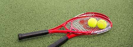 Two red tennis rackets lie overlapped, holding two yellow balls on their strings, on a green artificial-grass court.