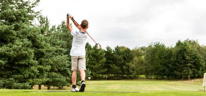 A child golfer swings a club overhead, finishing a drive on a tee box, with green fairway and tall pine trees under an overcast sky.