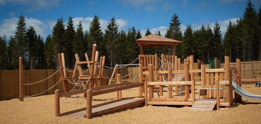 Wooden playground equipment forms interconnected climbing frames and rope bridges; a metal slide descends from a platform. Context: wood-chip ground, fenced perimeter, surrounding evergreen trees under blue sky.