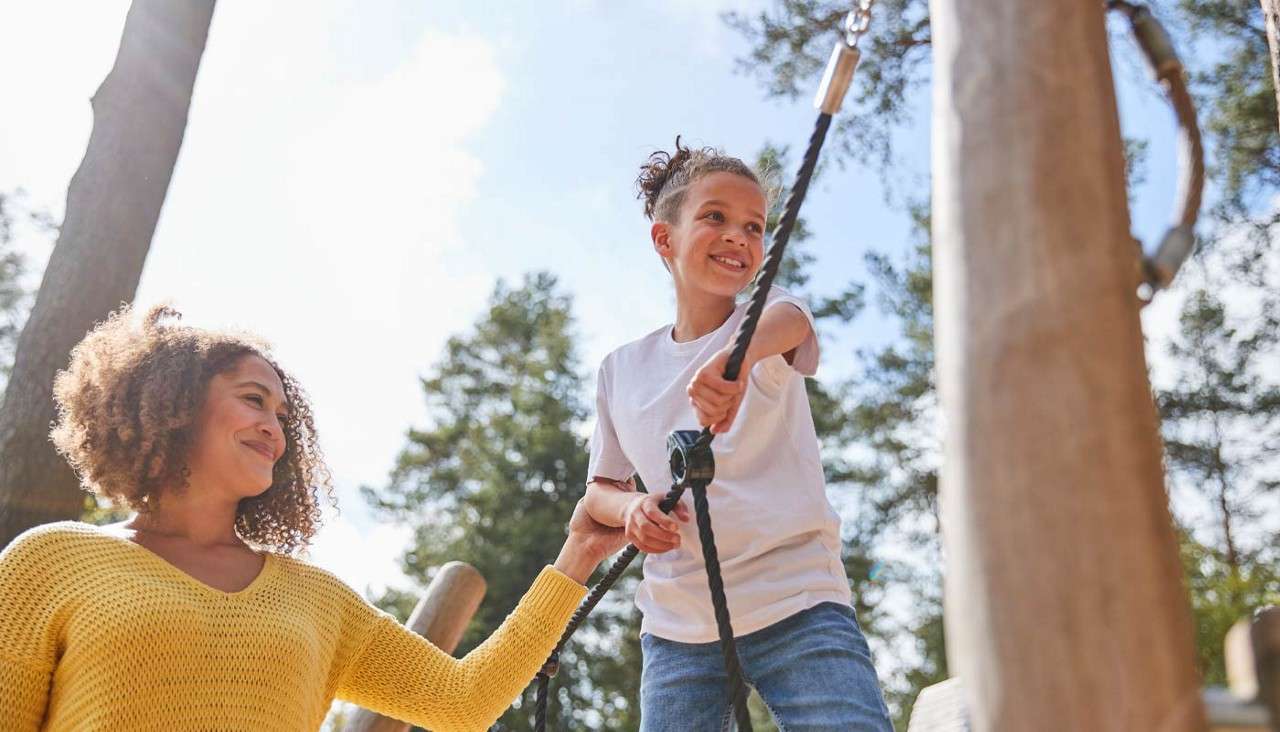 Child grips a rope zip-line handle, smiling while balancing. Adult beside them steadies their back and the rope. Wooden play structures and tall trees surround them on a sunny day.