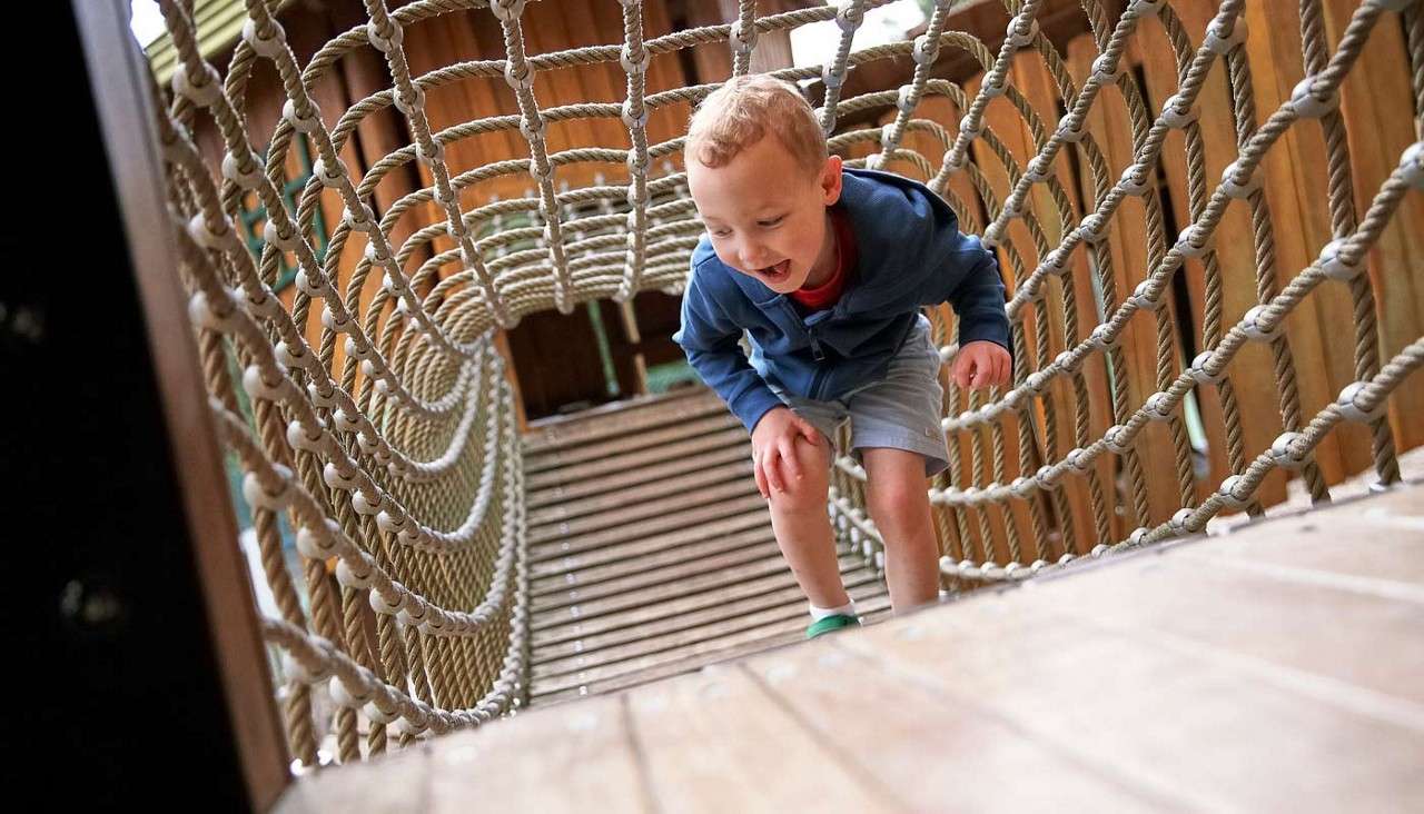 Child crouches and moves forward, smiling, on a netted rope tunnel bridge, surrounded by wooden planks and posts in an outdoor playground or treehouse structure.