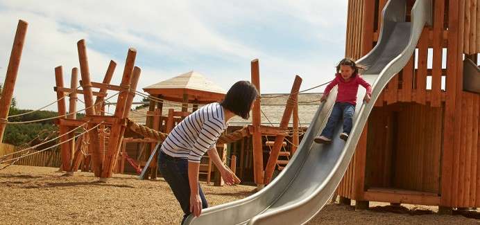 Child slides down a metal playground slide; adult stands beside, ready to catch; wooden climbing structures and ropes behind in a sunny outdoor play area.