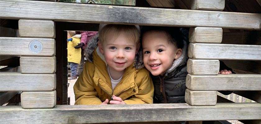 Two toddlers peer and smile through horizontal wooden slats, leaning on a play structure. Behind them, other children in coats move around in an outdoor playground under overcast light.
