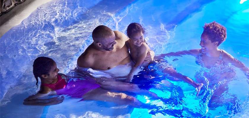 Four people laugh and play, sitting and standing in a brightly lit blue swimming pool at night, one adult holding a child while others relax against the shallow edge.
