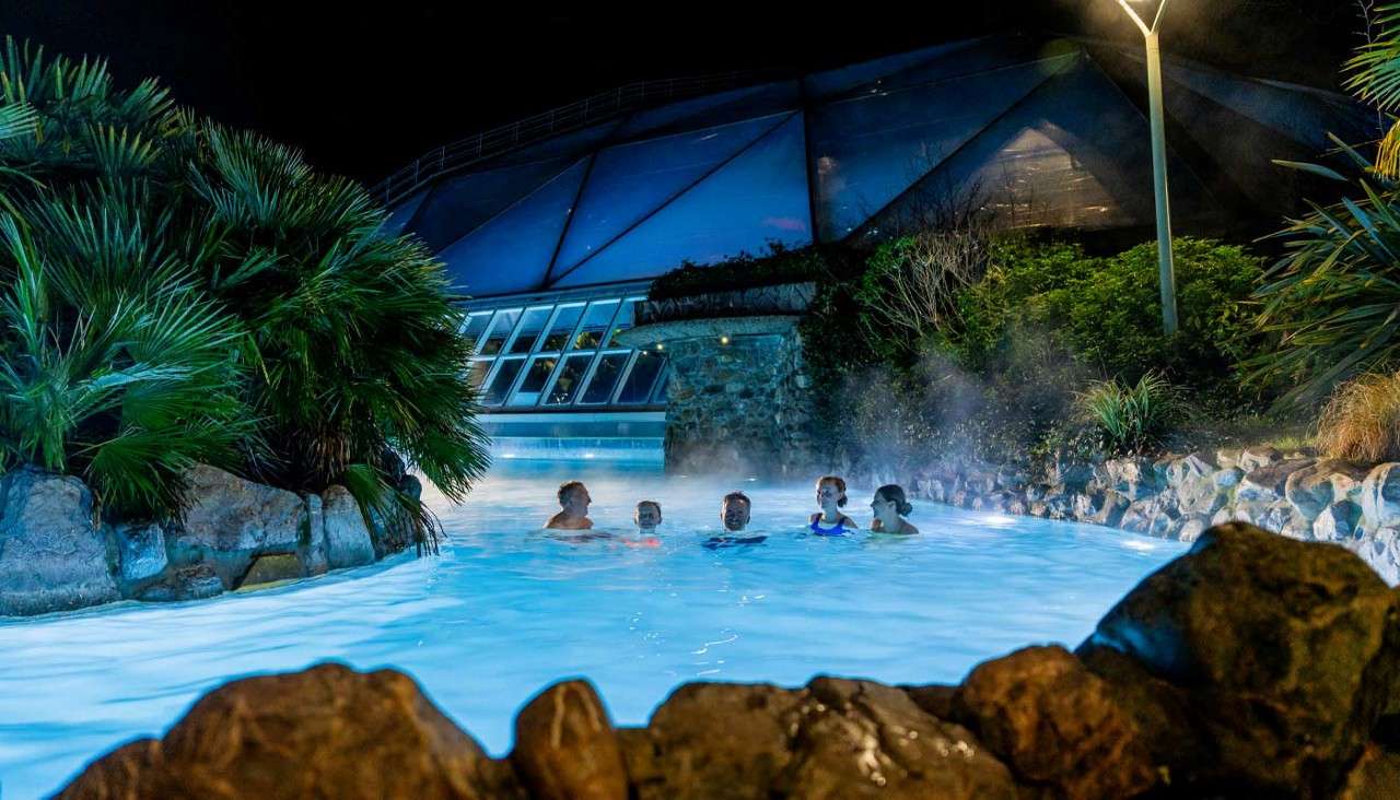 Swimmers relax and chat in a steaming, blue-lit outdoor pool, surrounded by rocks and tropical plants, with a futuristic glass-domed building and a lamppost glowing in the night.