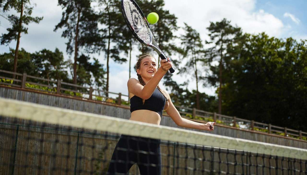 Tennis player swings a racket to hit a rising ball, smiling mid-forehand; net in foreground, outdoor court surrounded by tall trees under partly cloudy sky.