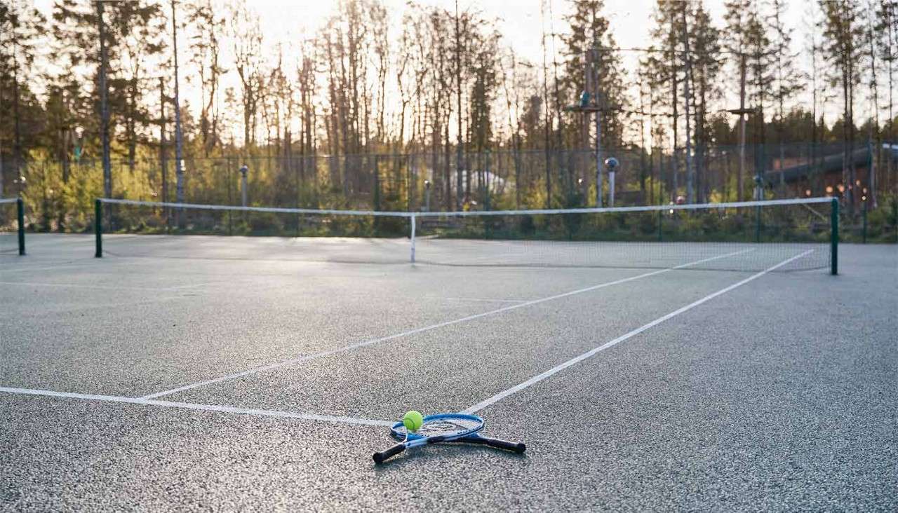 Tennis racket and ball resting on the court, lying near white lines; an empty doubles court stretches back with nets, surrounded by tall trees under soft evening light.